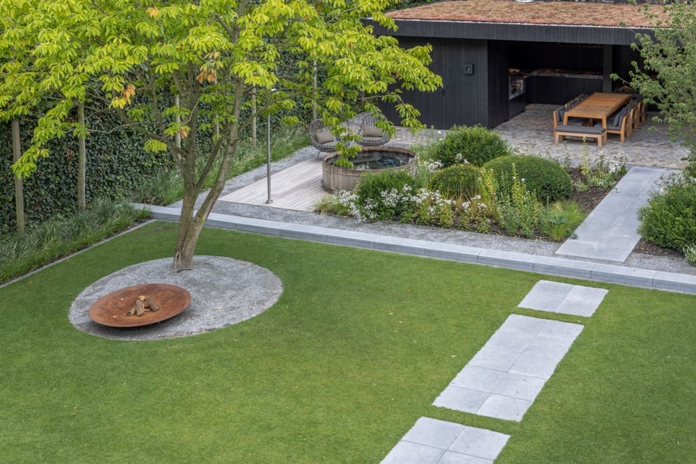 groot gazon met een pad er door heen en een cortenstaal vuurkorf onder de boom