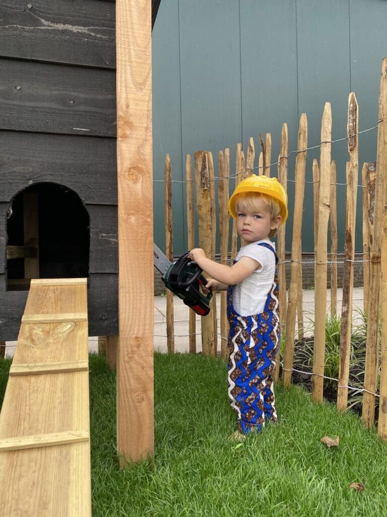 Peuter in tuinpak met bouwhelm speelt met speelgoedkettingzaag bij het kippenhok.