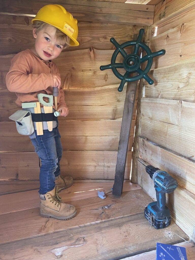 Peuter met gereedschap en bouwhelm helpt mee met timmeren in houten bouwwerk.
