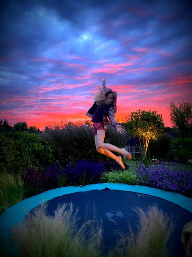 Meisje springt hoog in de lucht op Berg trampoline met ondergaande zon te midden van bloeiende tuin.
